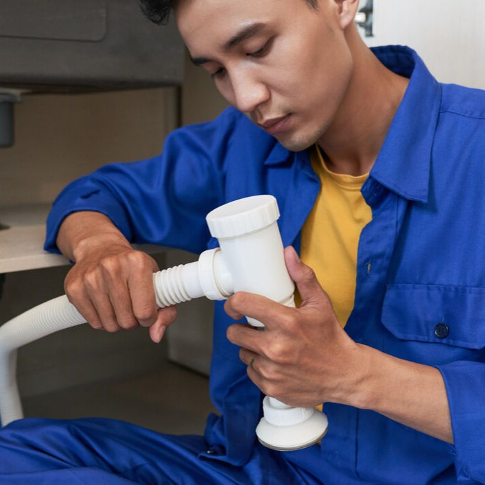 Man in blue coveralls repairing pipe under sink, focused expression. Indoor setting with beige background. Wearing yellow shirt.