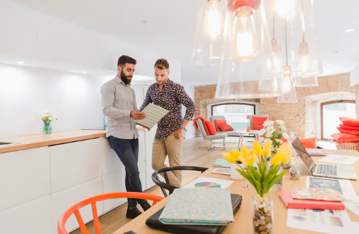Two men in a bright office review documents by a countertop. Table holds a laptop, colorful files, and tulips. Warm, modern decor.
