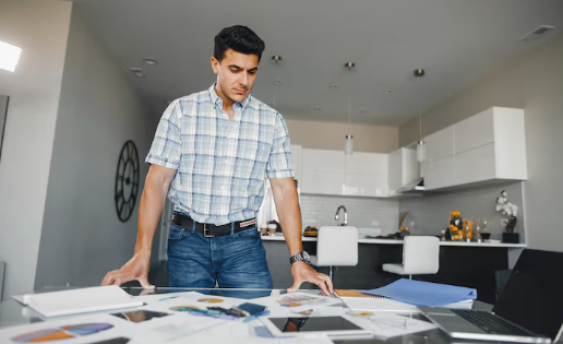 Man reviewing documents on table in modern kitchen, wearing plaid shirt and jeans. Bright, organized space with laptop and papers.