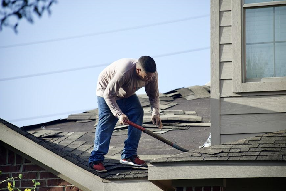Person in a beige shirt and jeans repairing a roof with a tool. Shingles are scattered. Beige house exterior. Clear blue sky.