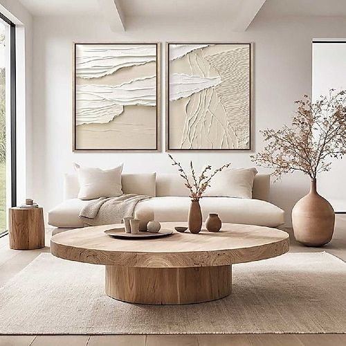 Minimalist living room with beige tones. Round wooden table, abstract wall art, vases with dried flowers. Bright and serene atmosphere.