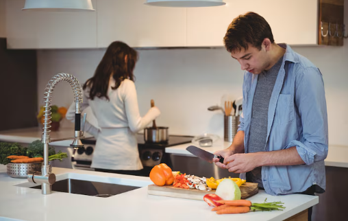 Man chopping vegetables; woman cooking at stove in modern kitchen. White cabinets, silver faucet, and colorful veggies create a cozy mood.