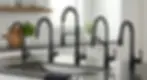 Four black kitchen faucets on a counter in a modern kitchen with white tiles and plants; sleek and minimalist design.