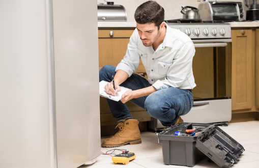 Man kneeling, writing on a clipboard near an open toolbox by a refrigerator in a kitchen. He looks focused. Kitchen utensils in the background.