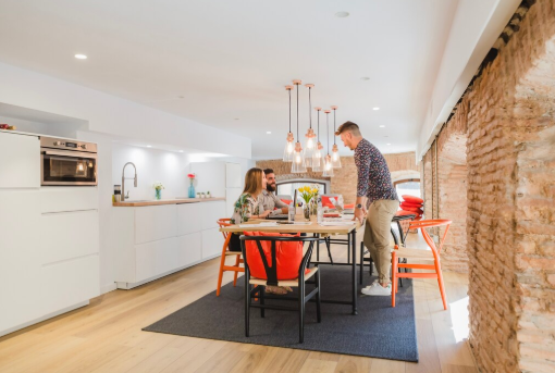 Three people sit and stand around a dining table in a modern kitchen with brick walls. Bright lights hang above, setting a cozy mood.