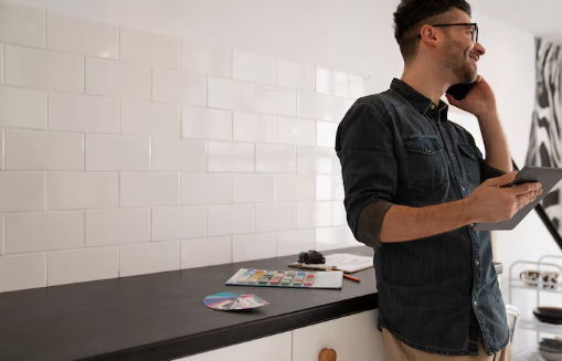 Man smiling, talks on phone holding tablet in modern kitchen with white tiles. Paint swatches and CDs on counter, zebra print on wall.