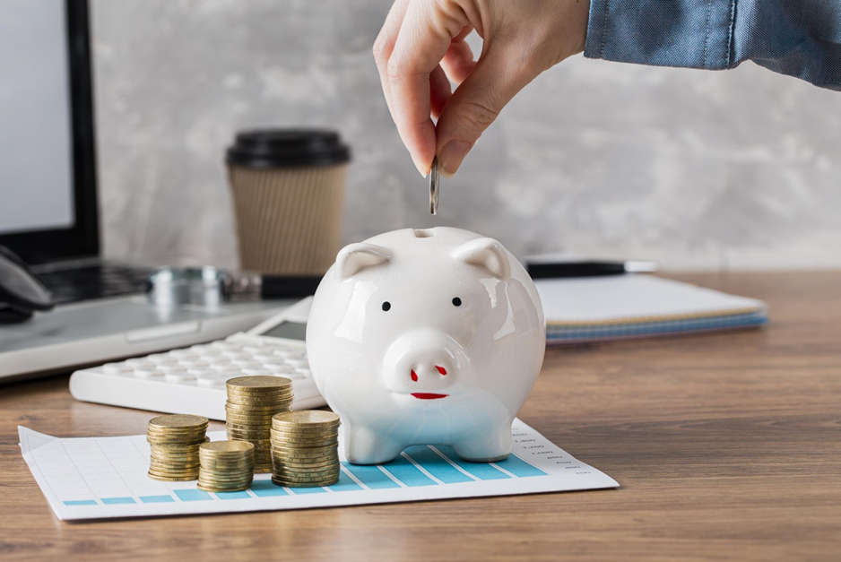 Hand placing coin in white piggy bank on a desk with stacked coins, a laptop, calculator, and a coffee cup in the background.