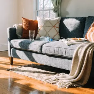 Gray sofa with orange and white pillows, a glass, water bottle, and plate on it. Sunlit room with wooden floors and green curtains. Cozy vibe.