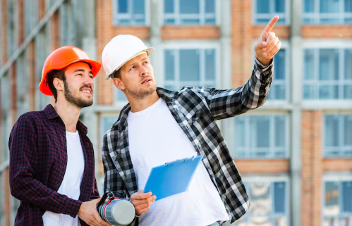 Two construction workers in helmets, one holding a blueprint, the other pointing, stand in front of a building, looking focused.
