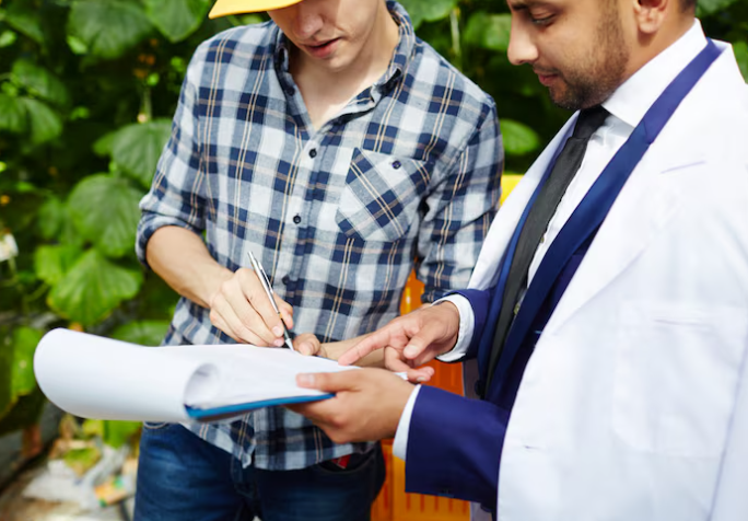Two men in a discussion; one in a plaid shirt taking notes, the other in a white lab coat pointing at a clipboard. Greenery in background.