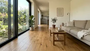 Bright living room with a beige sofa, wooden floor, potted plants, and large window showing a garden. Cozy and minimalist ambiance.