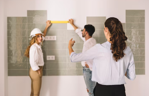 Three people in a room with light green tiles. One woman in a hard hat smiles, a man measures with a tape, and another woman gestures.