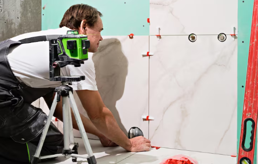 A man sets wall tiles using a level tool. He wears a white shirt and overalls. A green laser level on a tripod is in the foreground.