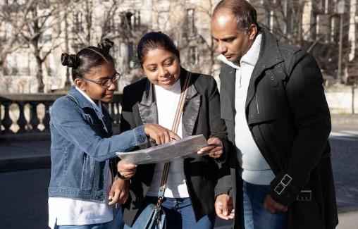 Three people study a map outdoors on a sunny day, standing on a street with bare trees in the background, displaying a focused mood.