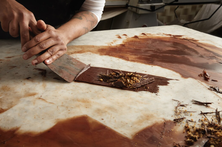 Hands shaping chocolate with a scraper on a marble surface. Chocolate shavings and crumbs scattered around. Warm, earthy tones.