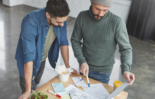 Two men reviewing documents on a wooden table with takeout food. One points at the papers. Room has gray flooring, casual setting.