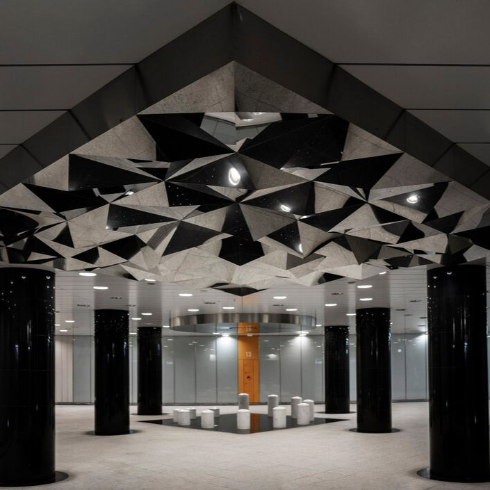 Geometric ceiling with black and grey patterns above a modern lobby setting. Black columns and white seating on the shiny floor. Moody lighting.