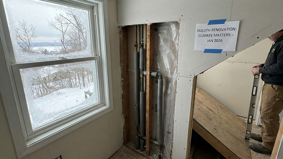 Window view of snowy landscape, pipes in wall under renovation, person holding a level. Sign reads "Duluth Renovation: Climate Matters – Jan 2026."