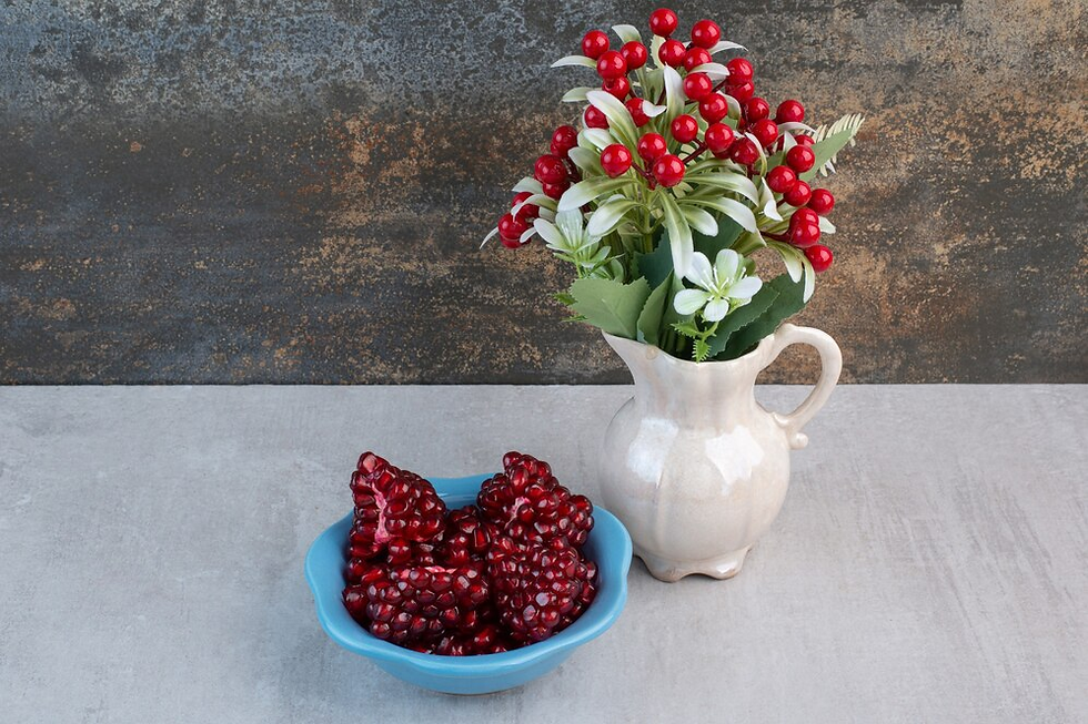White vase with red berries and green leaves on table next to blue bowl filled with pomegranate seeds. Rustic background.