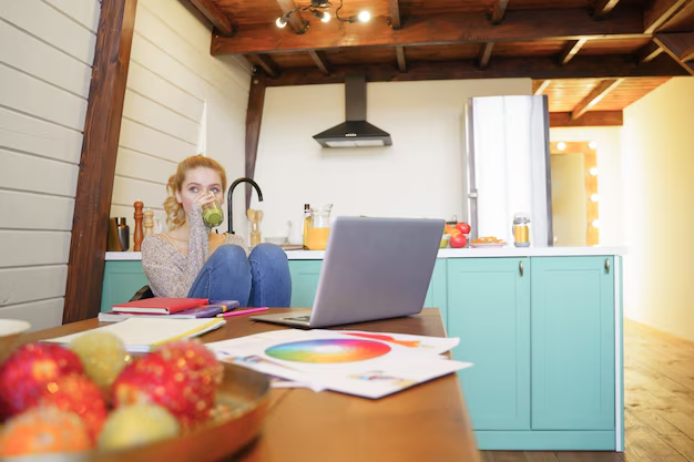 Woman sits in a cozy kitchen, sipping a drink by a turquoise counter. A laptop and color wheel printout are on the table. Warm lighting.