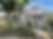 White house with a small porch, fenced yard, and hanging flower baskets. Sunny day with trees in the background. Peaceful suburban setting.