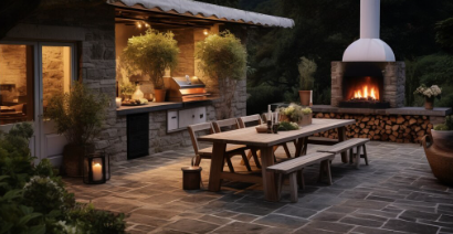 Outdoor patio with stone flooring features a wooden table, benches, and a lit fireplace. Warm lighting and potted plants create a cozy atmosphere.