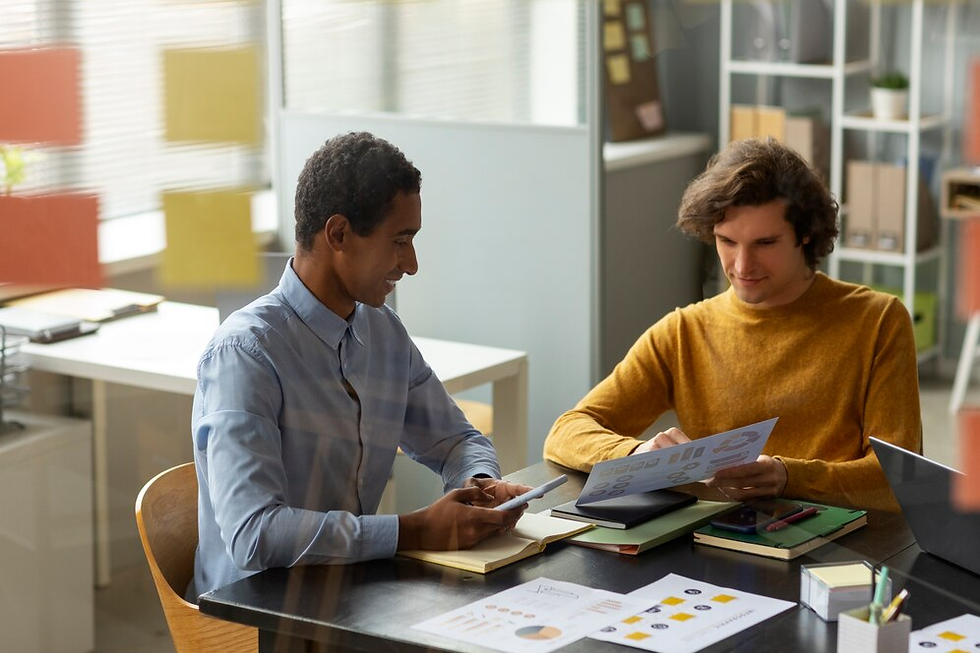 Two men at a desk reviewing a document with charts. One holds a tablet. Office setting with colorful sticky notes on windows. Relaxed mood.