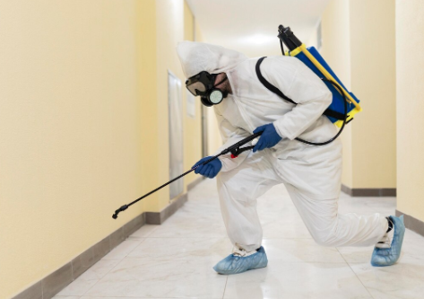 Person in white hazmat suit with blue gloves and mask sprays disinfectant in a beige hallway. Carries a blue and yellow backpack sprayer.