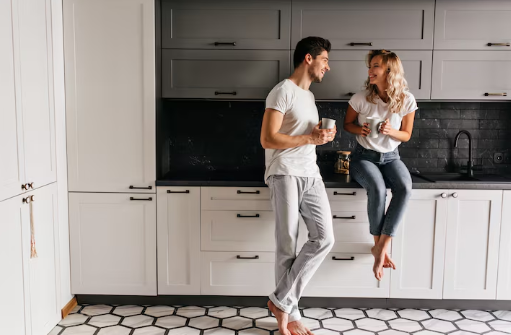 Couple in casual wear chatting in a modern kitchen with gray cabinets and hexagonal floor tiles, smiling and drinking coffee.