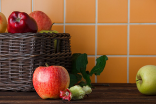 Basket with red and green apples on a wooden table, one apple with a bite. Orange tiled background, leafy greens beside.