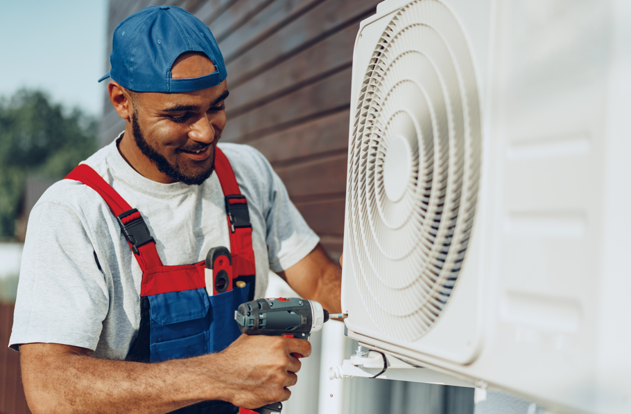 A man in a blue cap and red overalls smiles while using a drill to fix an outdoor AC unit. Background shows a wooden wall.