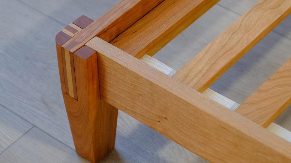 Wooden bed frame corner with dovetail joints on a light wood floor. The frame's warm wood tones contrast with the subtle background.