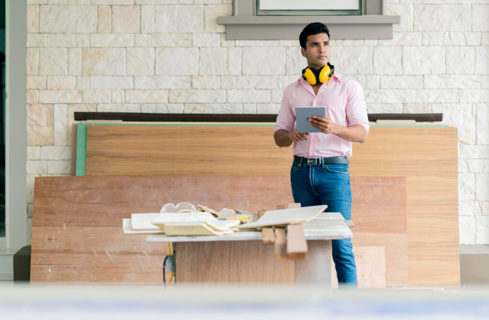 Man with yellow headphones holds a tablet, standing in a workshop with wooden panels and tools. Stone wall background. Contemplative mood.