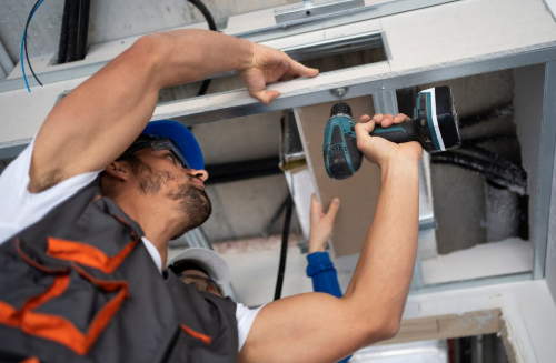 Workers in safety gear install overhead metal beams with a drill in a construction setting. Focused expressions. Gray and blue tones.