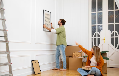 Man hangs frame on wall; woman on floor points up. Boxes and ladder nearby in a bright room. Casual mood, neutral colors.