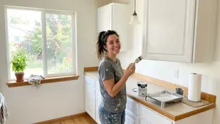 Woman painting kitchen cabinets white, smiling. Bright room with large window, visible garden. Paint can reads "White Satin". Relaxed mood.