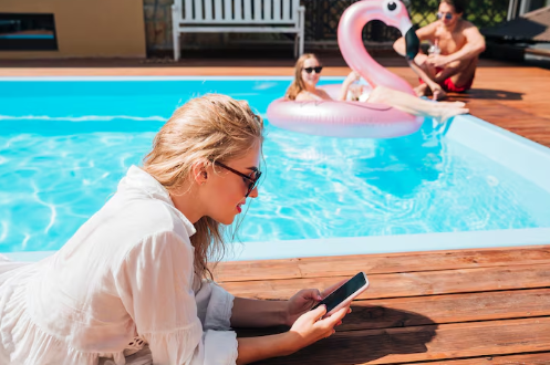 Woman in white shirt uses phone by a pool. Two people relax on a pink flamingo float. Sunny setting with wooden deck. Relaxed mood.