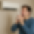 Man sneezing into tissue near wall-mounted air conditioner. Wearing a blue sweater, appearing uncomfortable in a beige room.