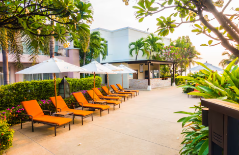 Poolside area with orange lounge chairs, white umbrellas, and lush greenery in a tropical setting. Bright, sunny atmosphere.