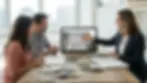 Three people discuss samples at a wooden table in a modern office. A screen displays a home decor site. Bright natural light enters from windows.