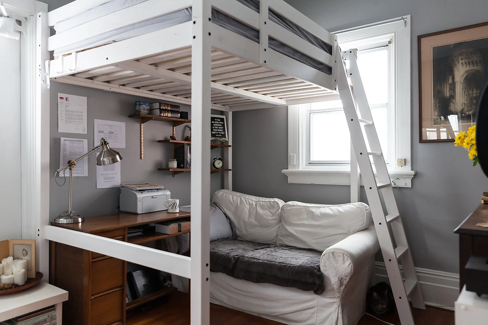 Loft bed with ladder over a cozy workspace featuring a desk, lamp, printer, and white sofa. Gray walls, window, and flower vase visible.