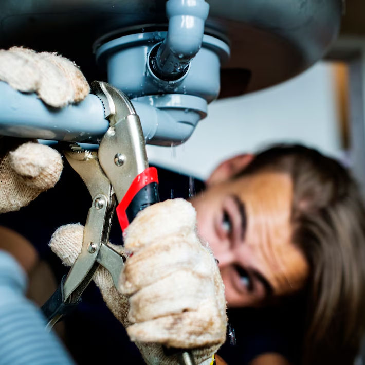 Person tightening a leaking pipe with a wrench under a sink. Wearing gloves. Focused expression. Bright lighting, close-up view.