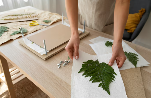 Hands pressing green fern leaves in a workshop setting. Screws, a wooden press, and yellow scissors are on a light wood table. Bright, cozy mood.