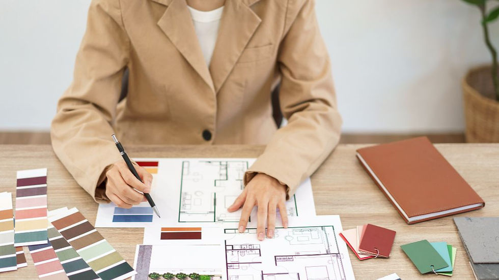 A person in a tan blazer works at a desk with architectural plans, color swatches, and a notebook, showcasing a design process.