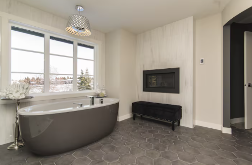 Modern bathroom with gray hexagonal tiles, a freestanding tub, chandelier, and wall fireplace. Large window shows a snowy landscape.