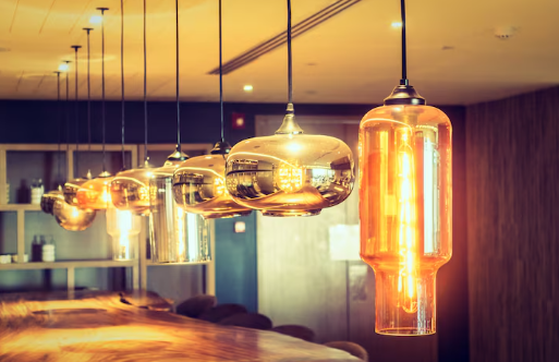 Vintage pendant lights with warm glow over a wooden table in a cozy, modern interior. Background features shelves and soft lighting.