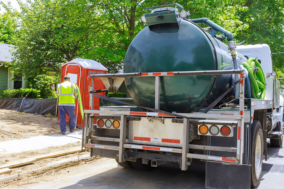 Worker in a yellow vest services a red portable toilet with a green hose from a septic truck. Trees and a house are in the background.