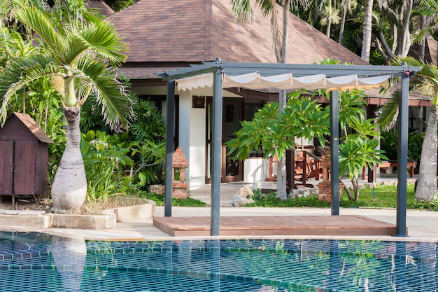 Tropical setting featuring a blue-tiled pool, pergola, lush green plants, and a wooden-roofed building. Calm and serene atmosphere.