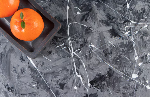 Two oranges with leaves in a dark tray on a splattered black and gray abstract background, creating a vibrant and artistic contrast.