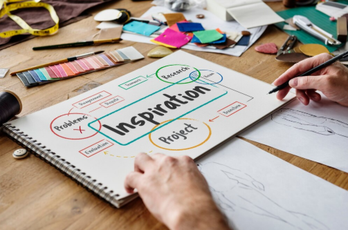Hands drawing on a sketchbook labeled "Inspiration" surrounded by colorful stationery on a wooden table, evoking creativity and planning.